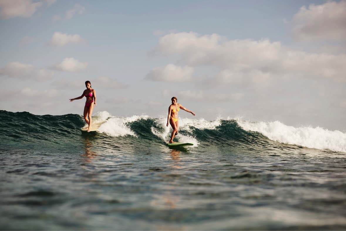 Two surfers riding a small clean wave on longboards in the Maldives under a tropical sky