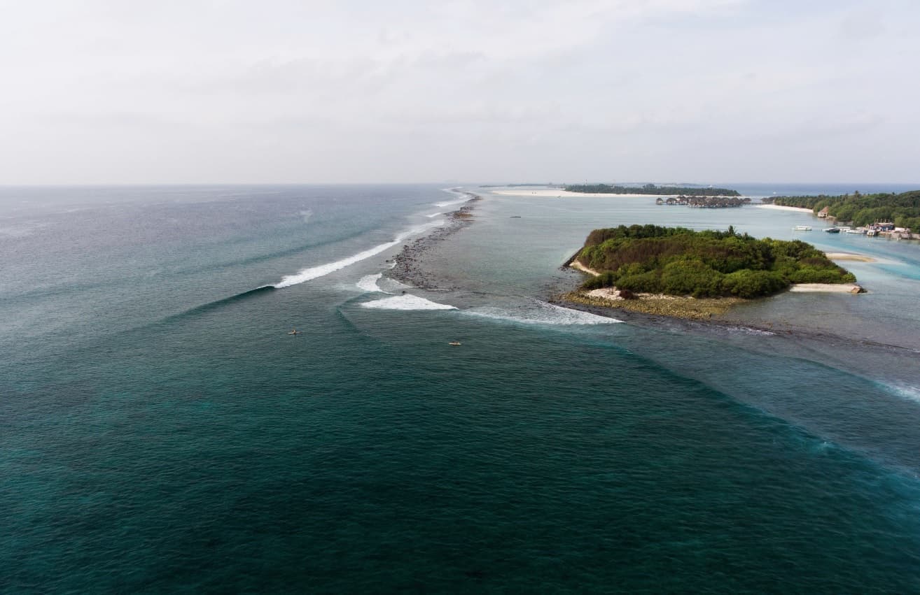 Aerial drone view of a Maldives surf pass with waves wrapping along the reef and surfers in the lineup near a tropical island