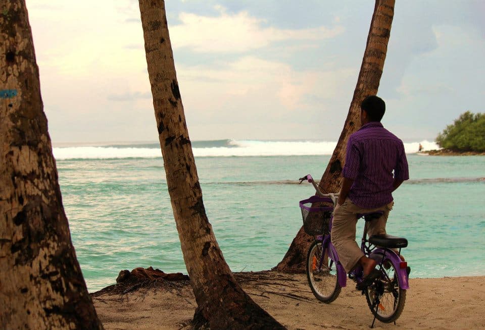 Local man sitting on a bicycle watching waves break on a reef from a tropical beach in the Maldives